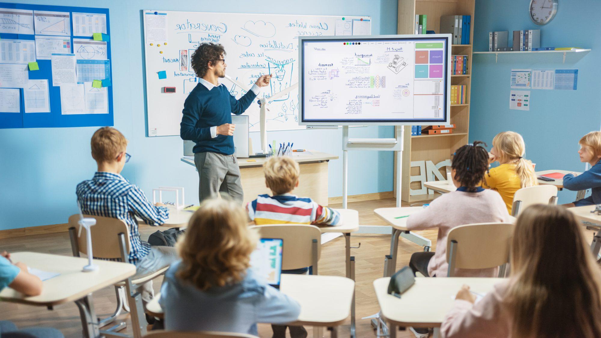 Man pointing to a white board in front of elementary students in desks