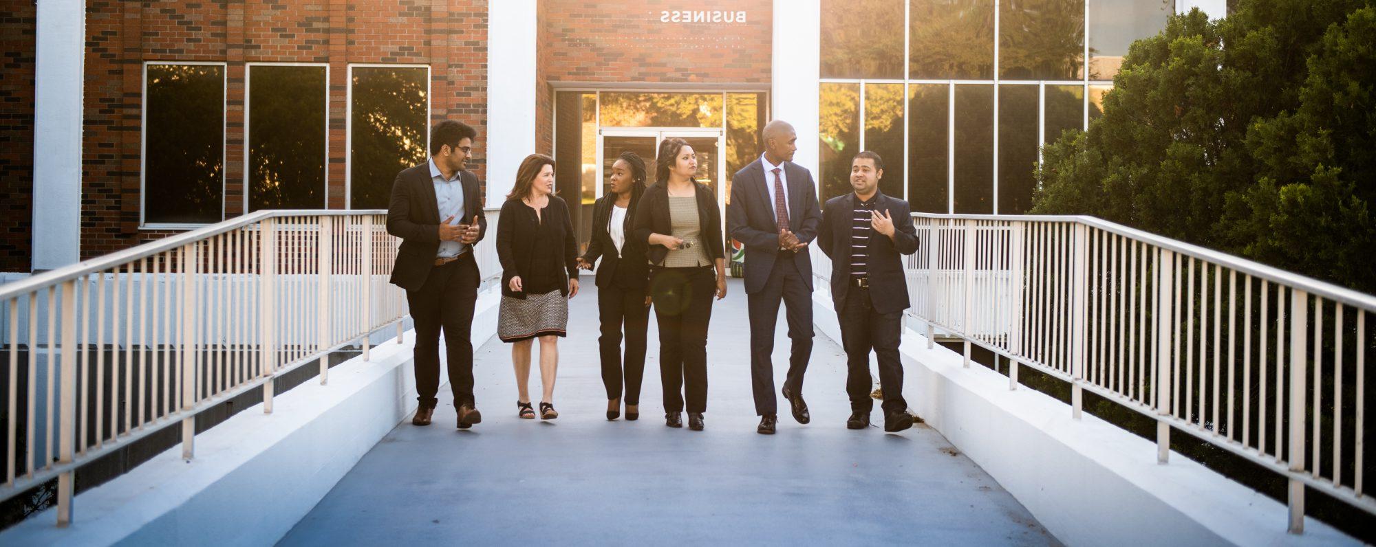 A group of business students talking to eachother while walking outside the business building.