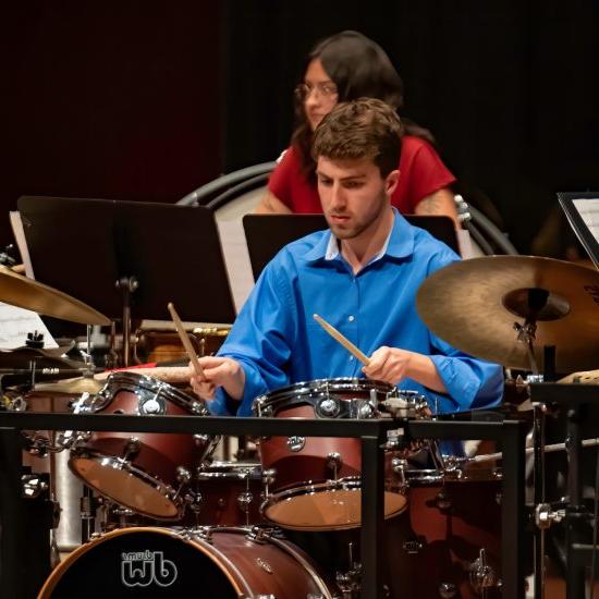 A person plays a drumset while another person plays a percussion instrument in the background.