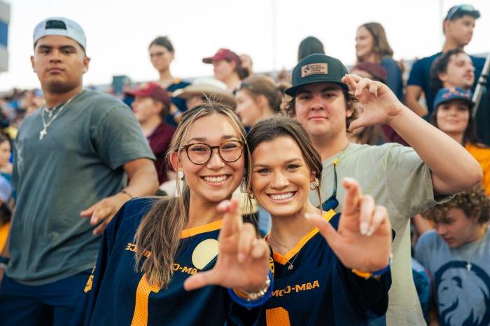 学生 in bleachers at football game smile into camera and flash the Lucky sign (fingers crossed).