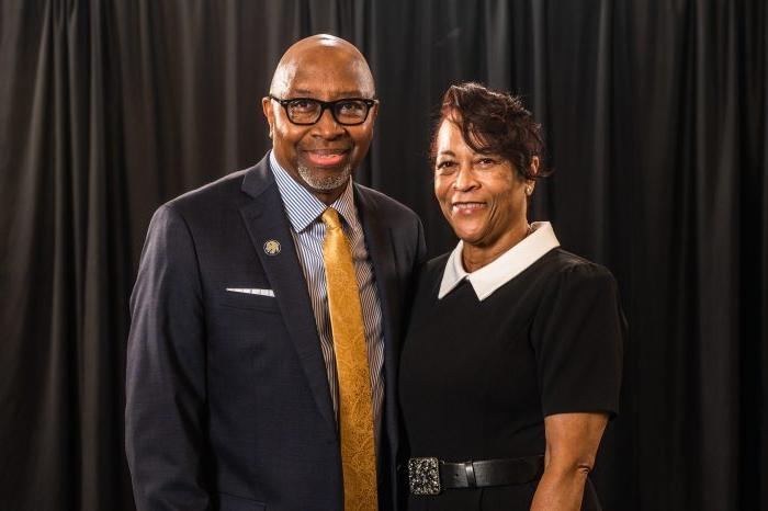 Derryle and Carolyn Peace face the camera and smile. Couple is dressed formally with black curtained background.