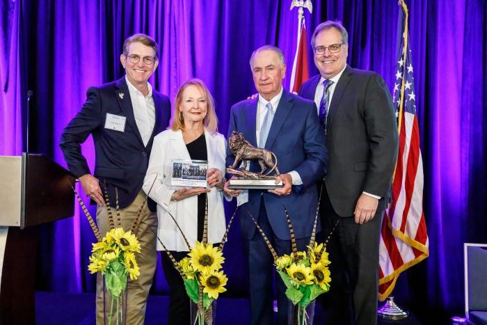 From left to right, Dr. Rudin, Senator Ted Lyon, Donna Lyon and Dr. Kelly 瑞娜 stand on stage in front of a purple curtain and the U.S. 和德州旗帜. The Lyons are holding a lion statue. In front of the people, three bouquets of sunflowers sit on the floor.