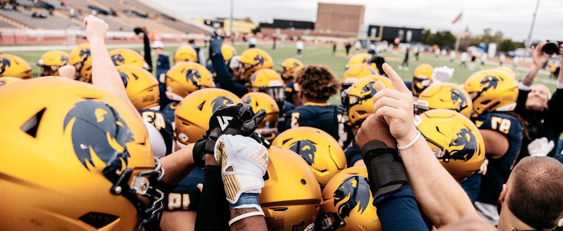 足球运动员在进场前排好队. Photo shows the tops of several players' helmets; some players have their arms raised. 背景是足球场.