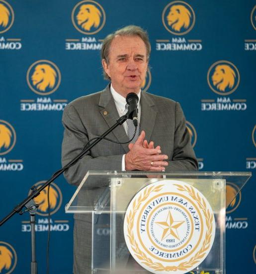 Chancellor John Sharp speaks behind a podium with hands clasped. A blue backdrop with the A&M-Commerce logo is behind him.